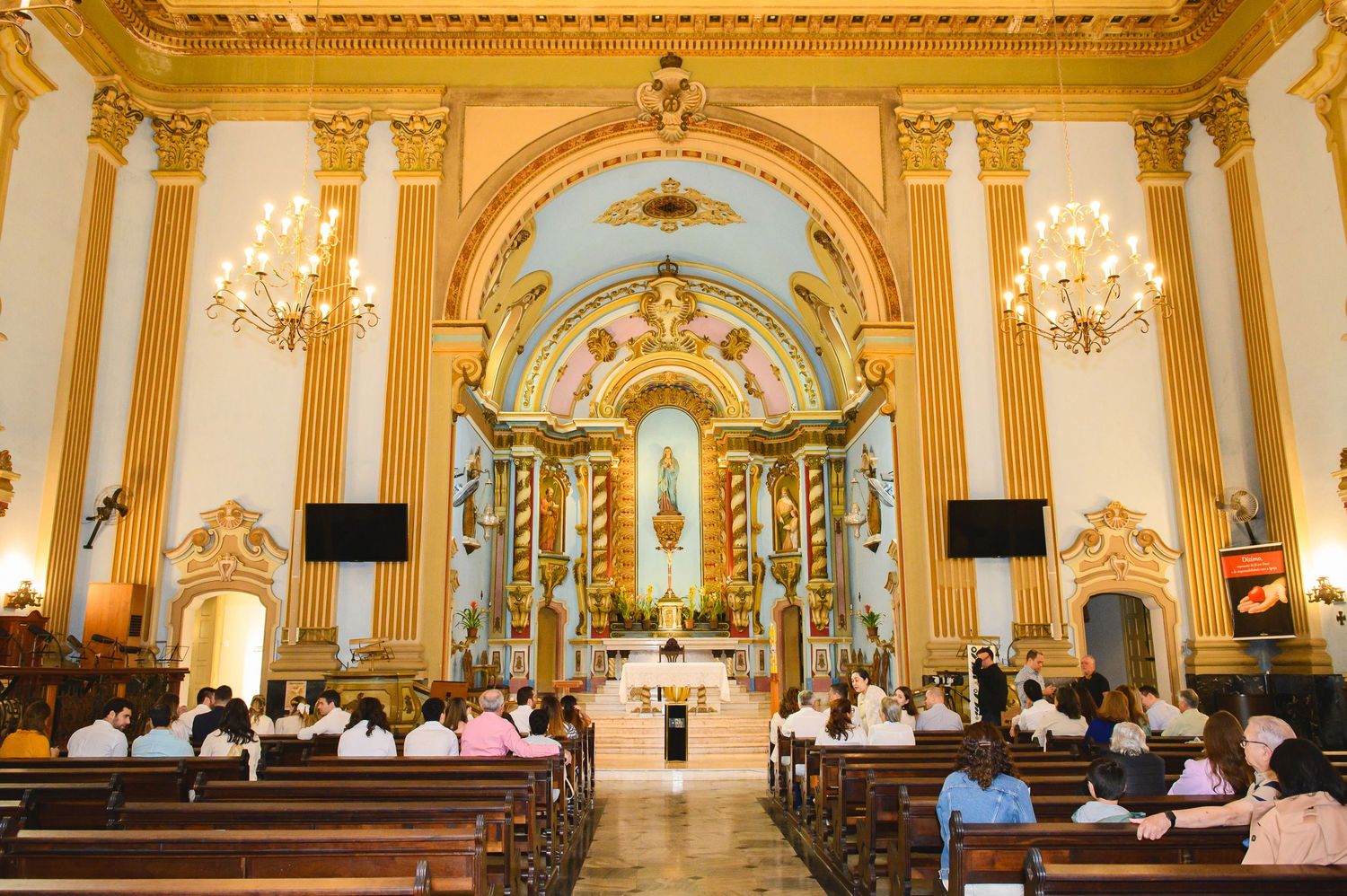 Interior of an ornate Catholic church with golden columns, crystal chandeliers and gathered congregation during mass.