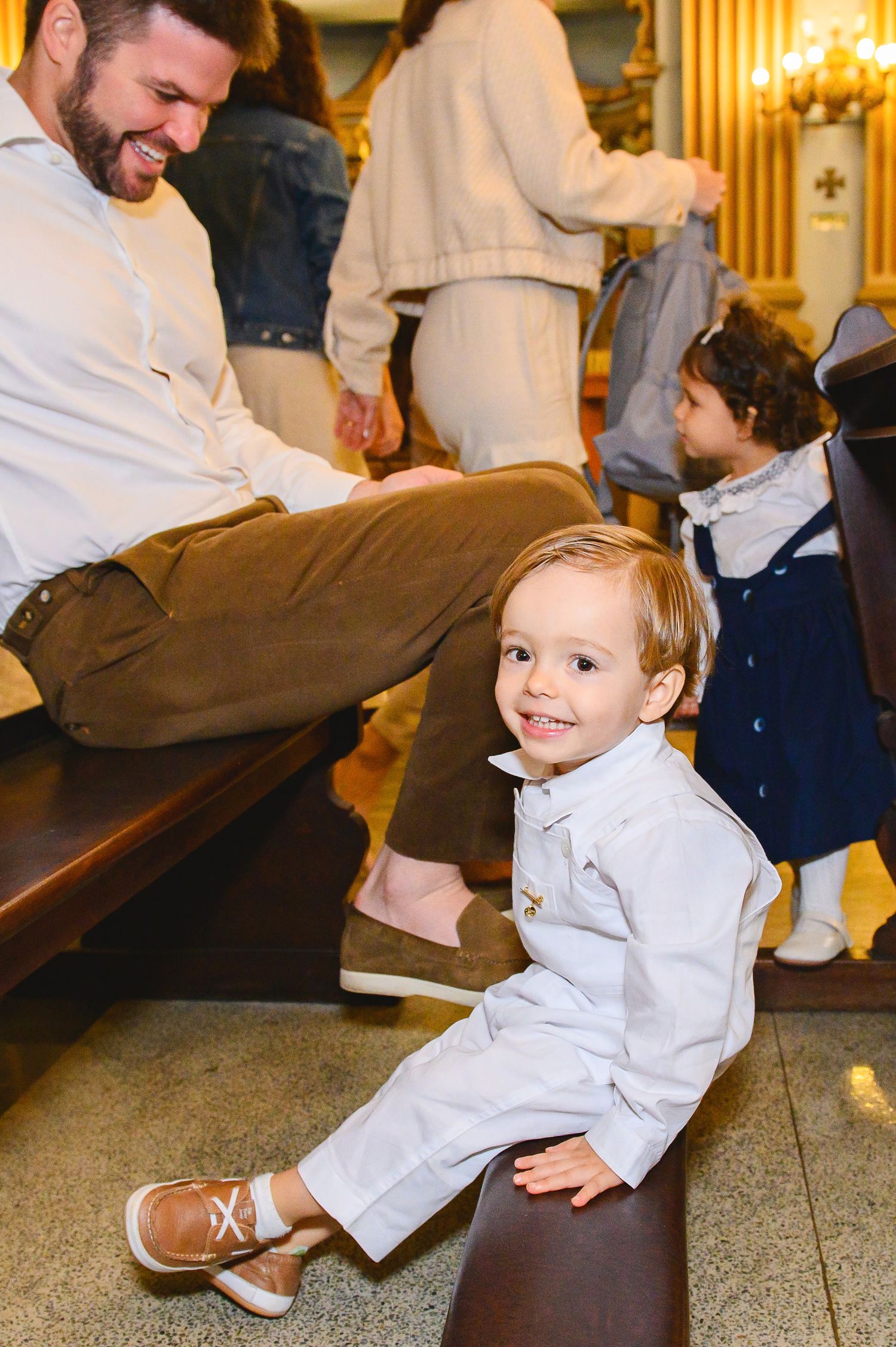 A toddler wearing a white outfit sits on a wooden church pew with a cheerful expression during a gathering.