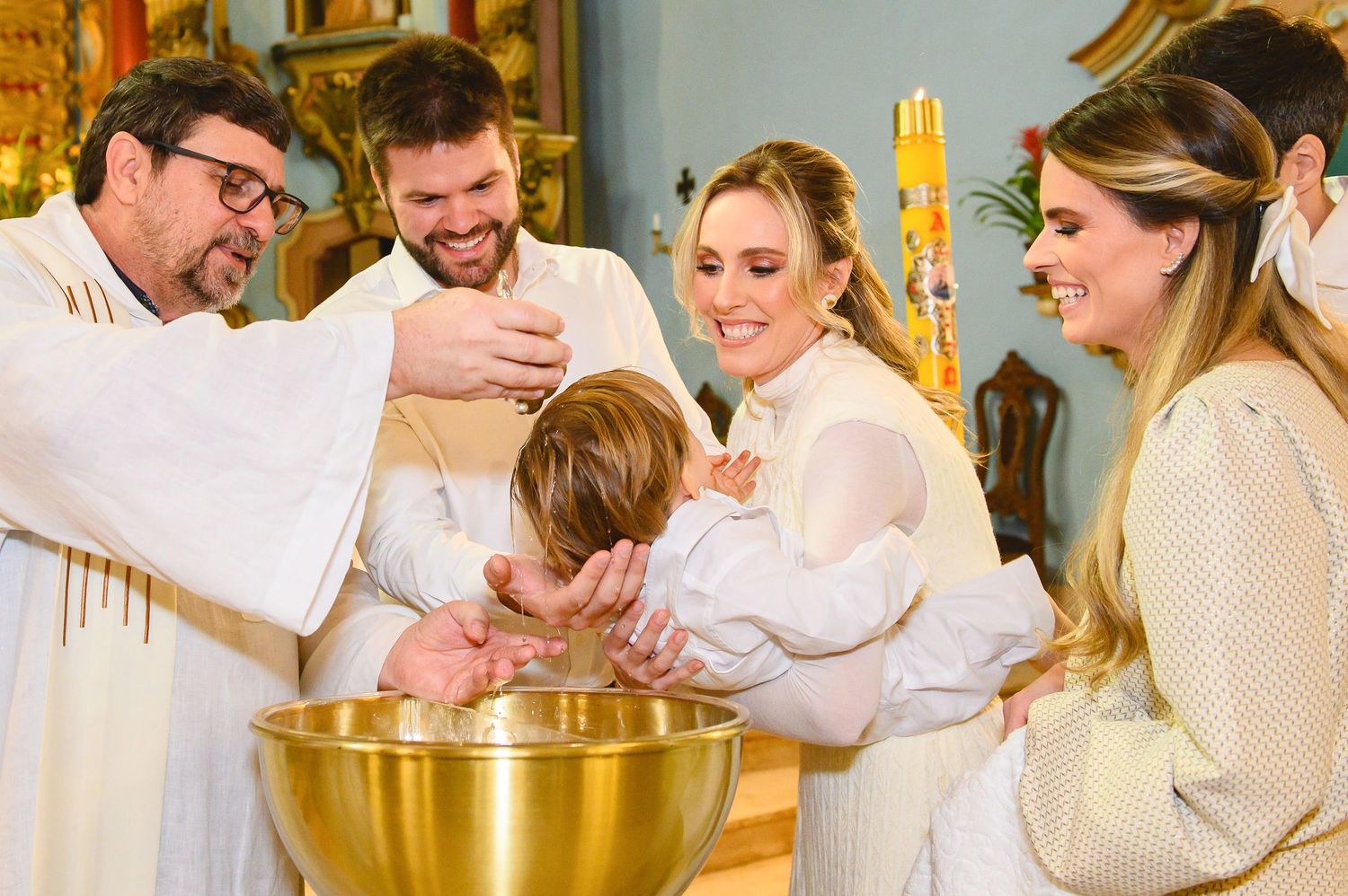 A priest performs a baptism ceremony over a golden font while family members gather around during the religious sacrament.