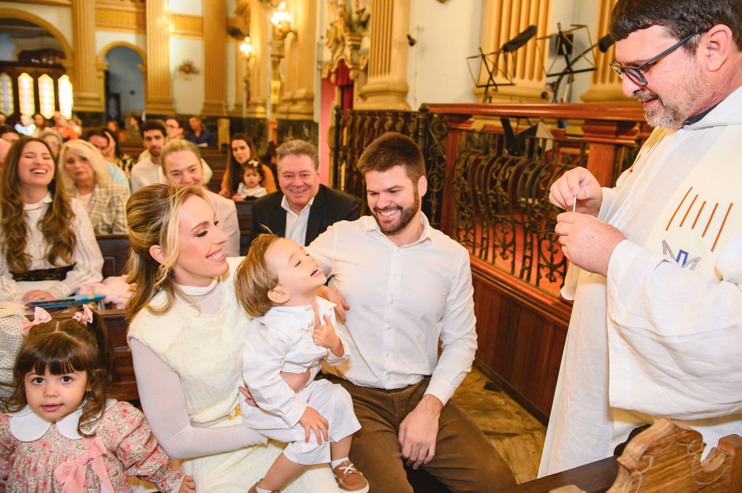Joyful baptism ceremony inside an ornate church with family and priest gathered around a baby in white.