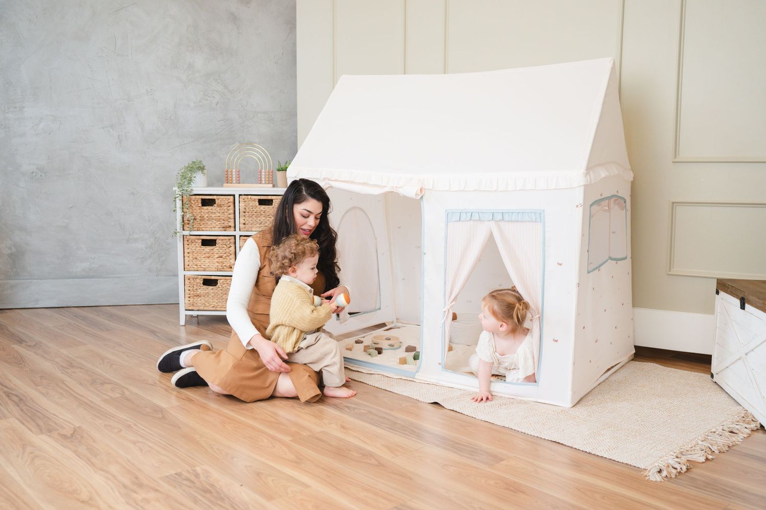 Two people sit together playing next to a white indoor fabric playhouse tent on a light wooden floor.