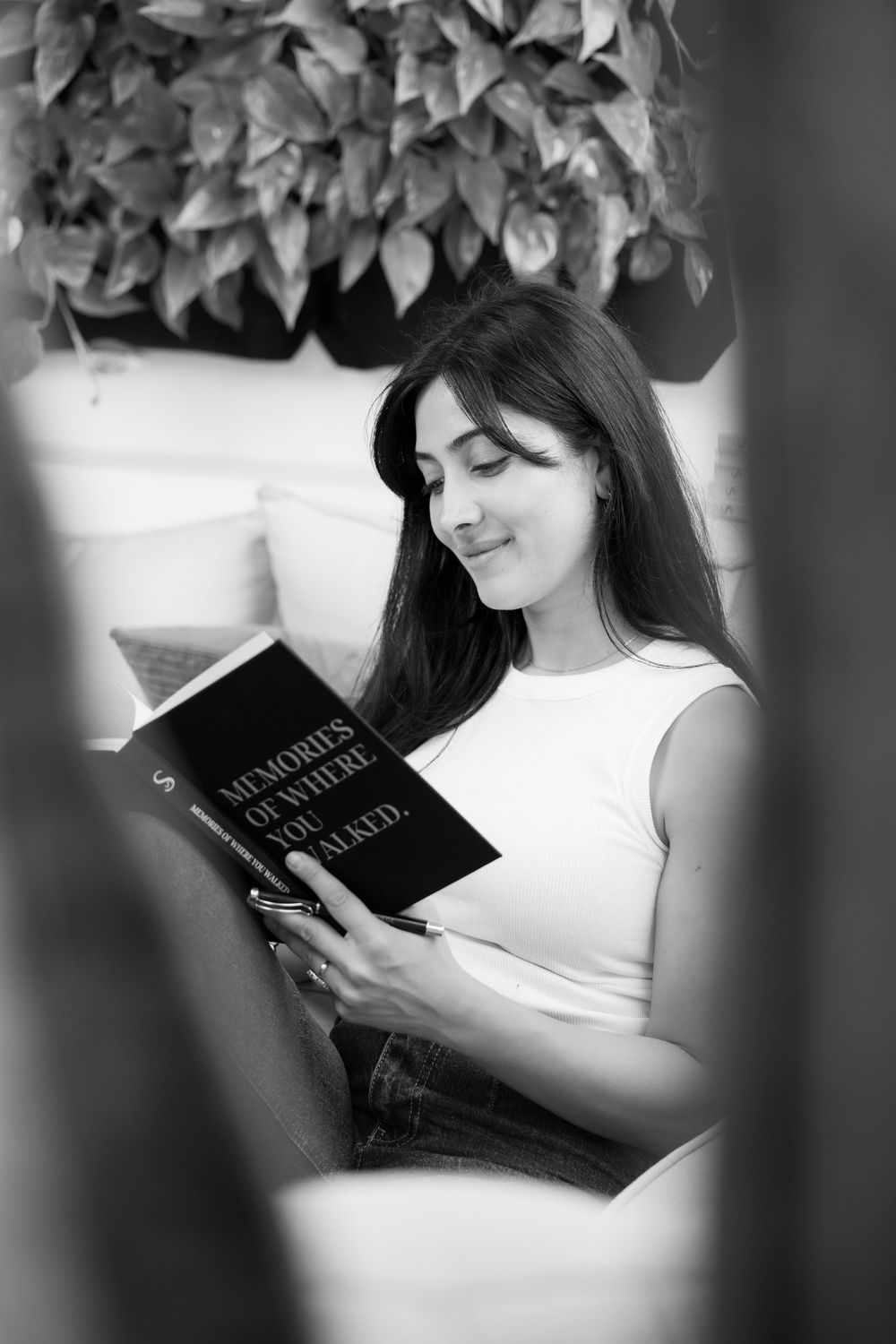 Black and white photo of woman reading book while seated on couch under indoor plants.