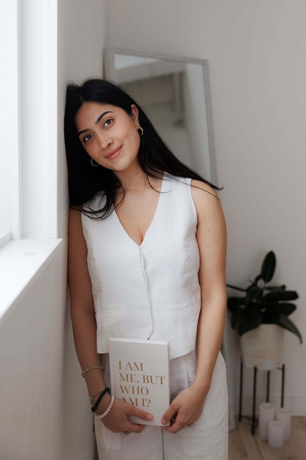 Woman in white dress holding book by window in minimalist interior setting.