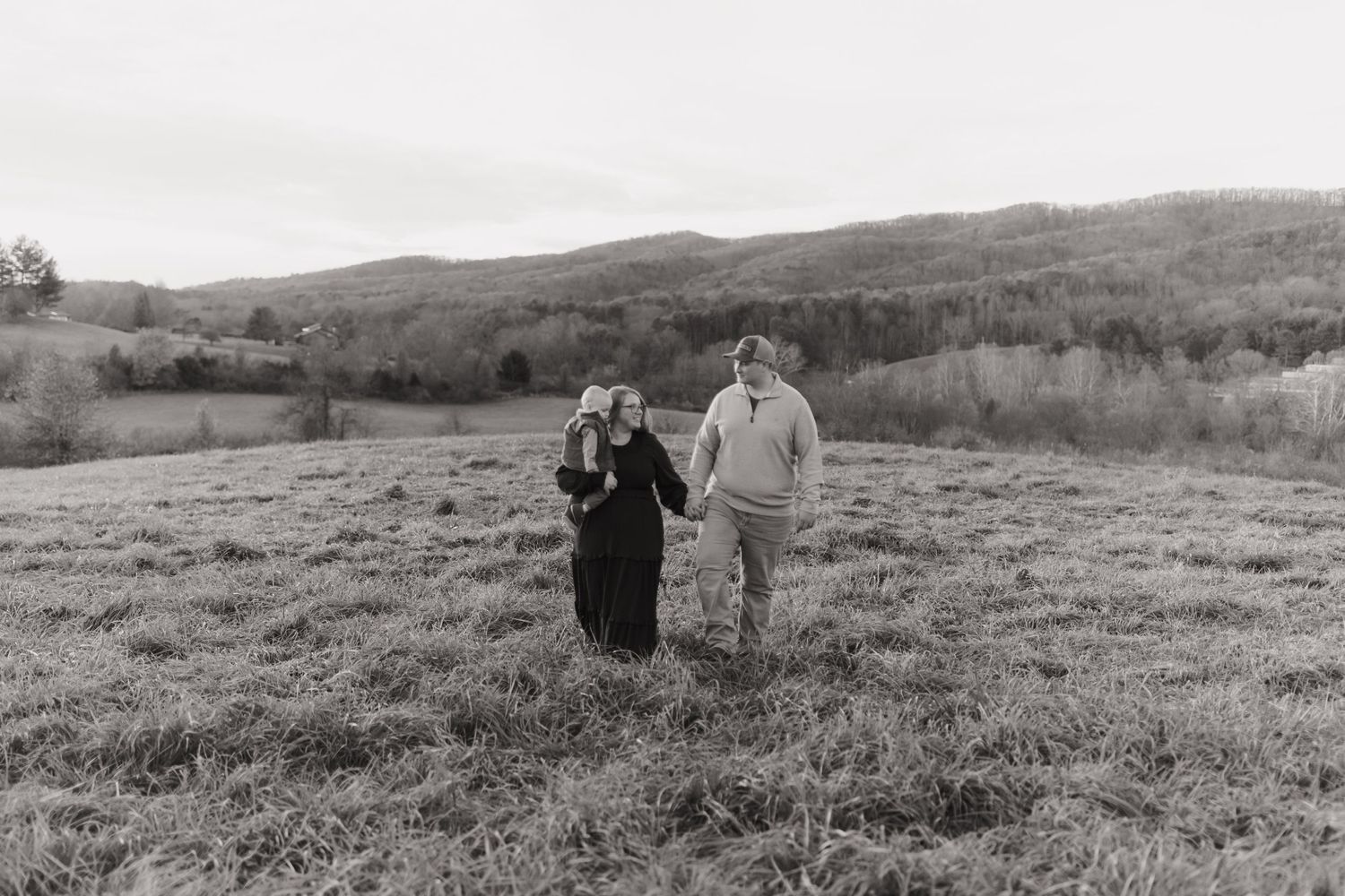 Black and white photo of couple walking together across open field with mountains in background.
