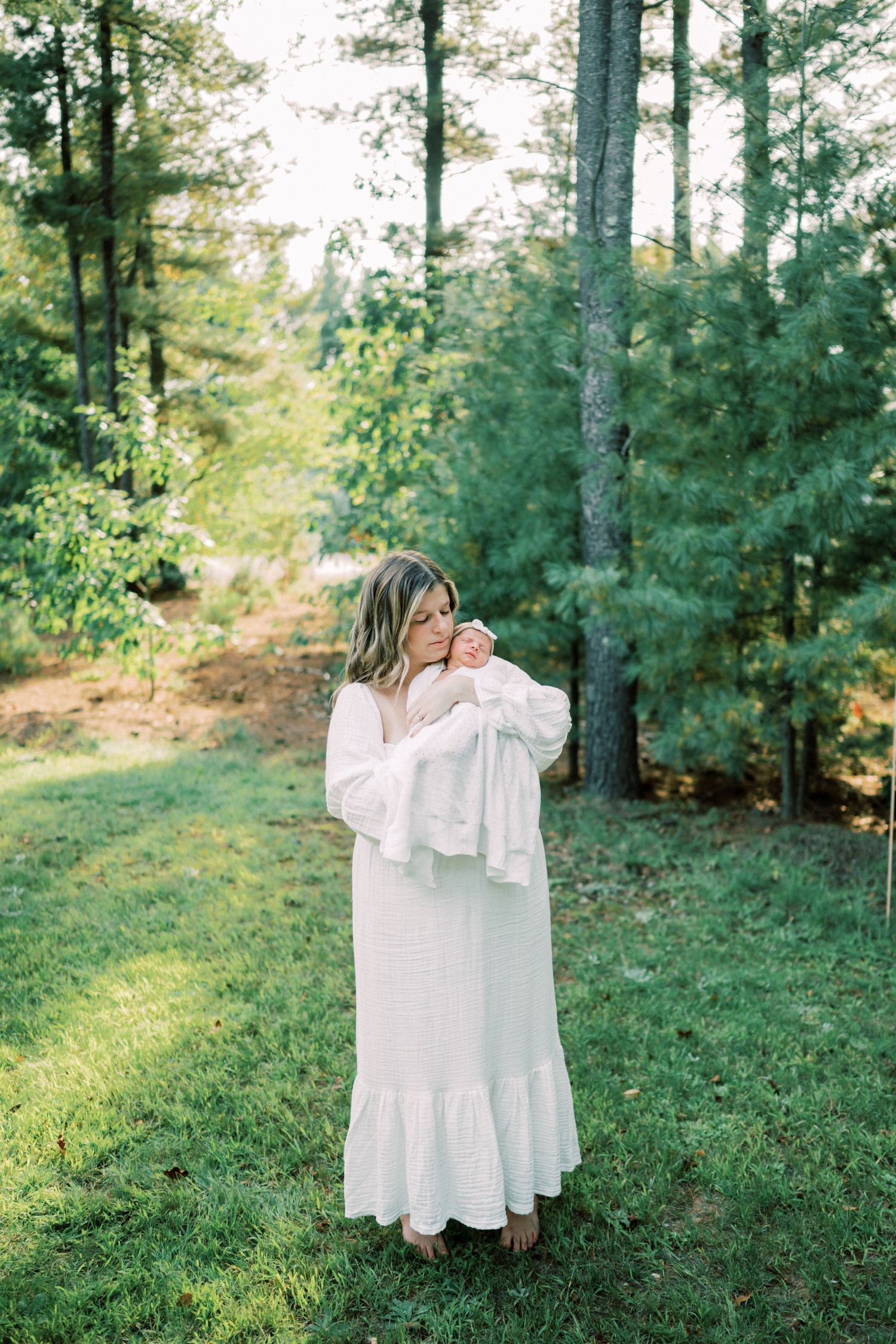 Woman in flowing white dress holds baby while standing in sunlit forest setting.
