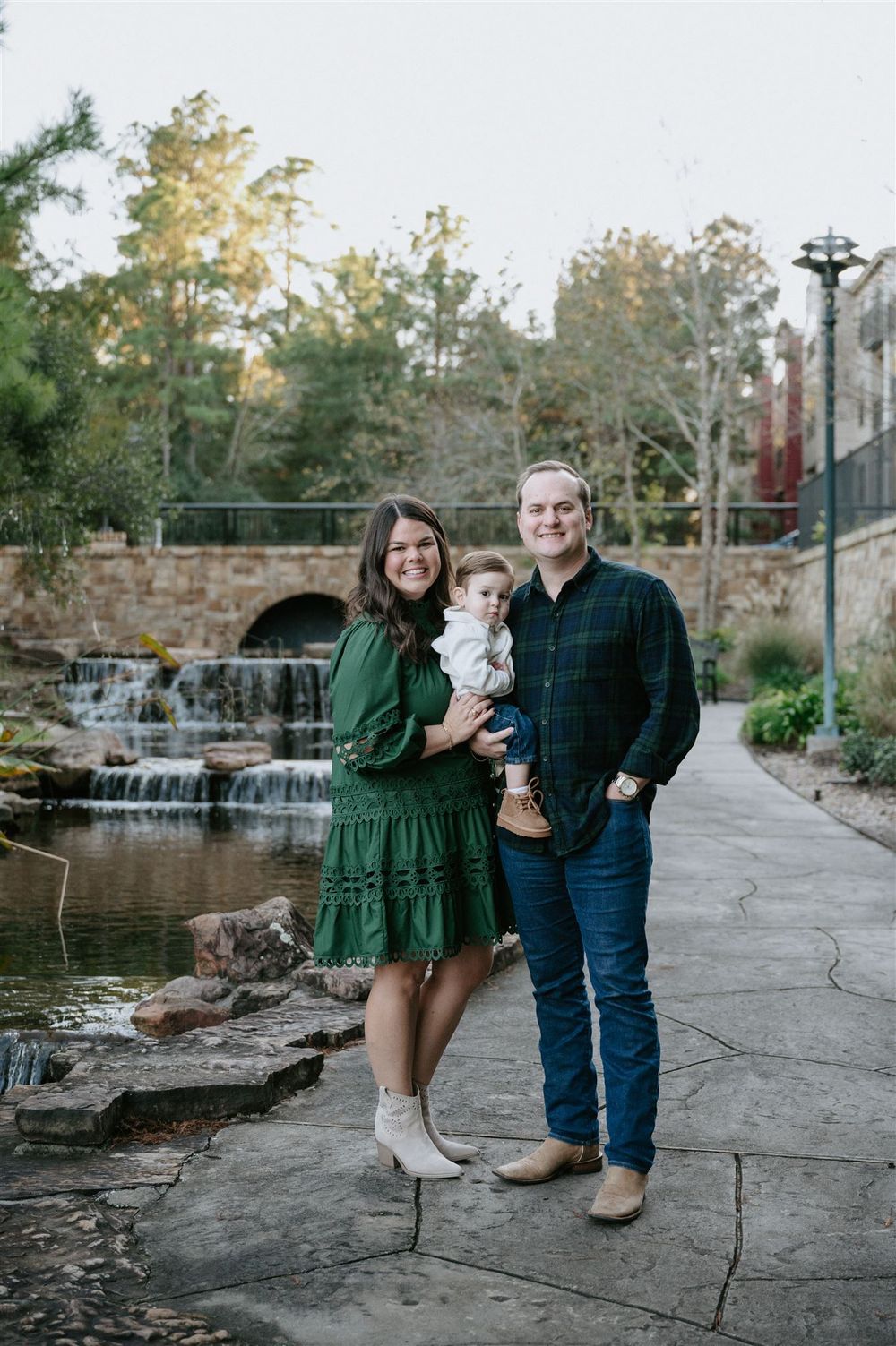 Family stands together on stone pathway near cascading waterfall and autumn trees in outdoor park setting.