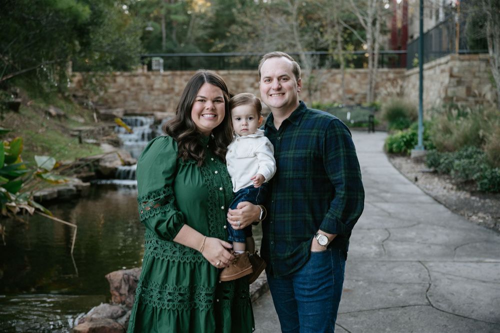 Three people in coordinating green and navy outfits pose together on a paved path next to a peaceful stream.