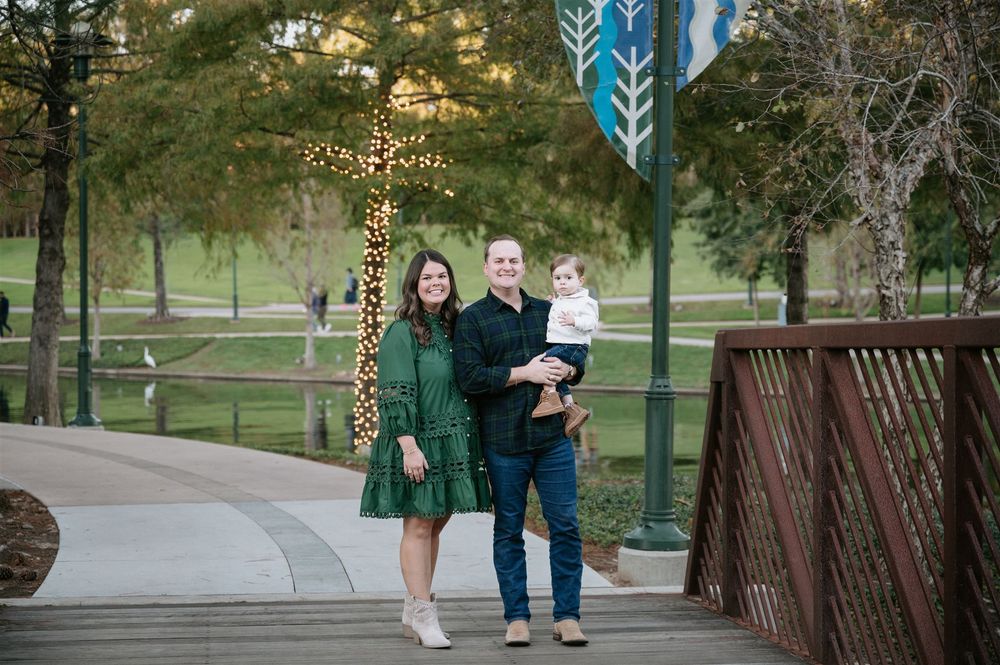 A family stands on a wooden bridge surrounded by illuminated holiday decorations and trees in a park at dusk.