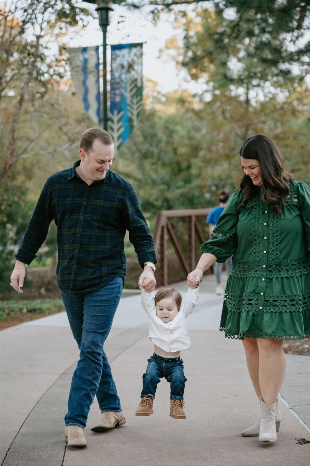 A family of three walk together on a pathway in a park, with the parents supporting a toddler's first steps.