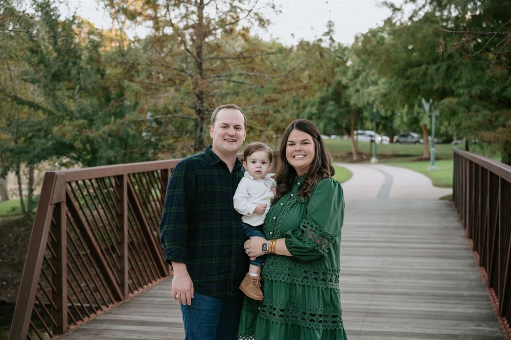 A family poses together on a wooden bridge surrounded by green trees in a park setting during golden hour.