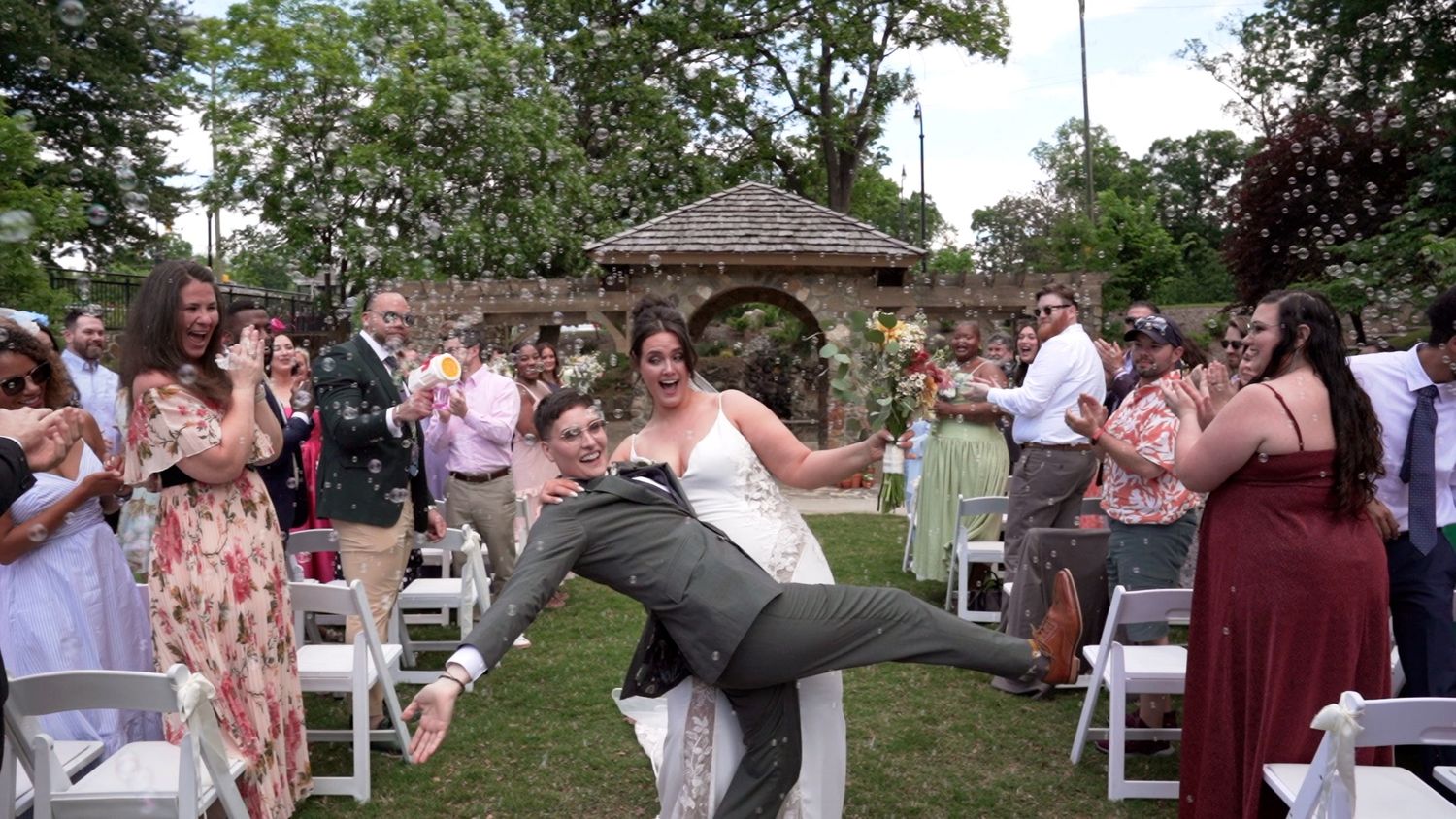 A couple performs a dramatic dip during an outdoor garden wedding celebration as guests cheer and watch from white chairs.