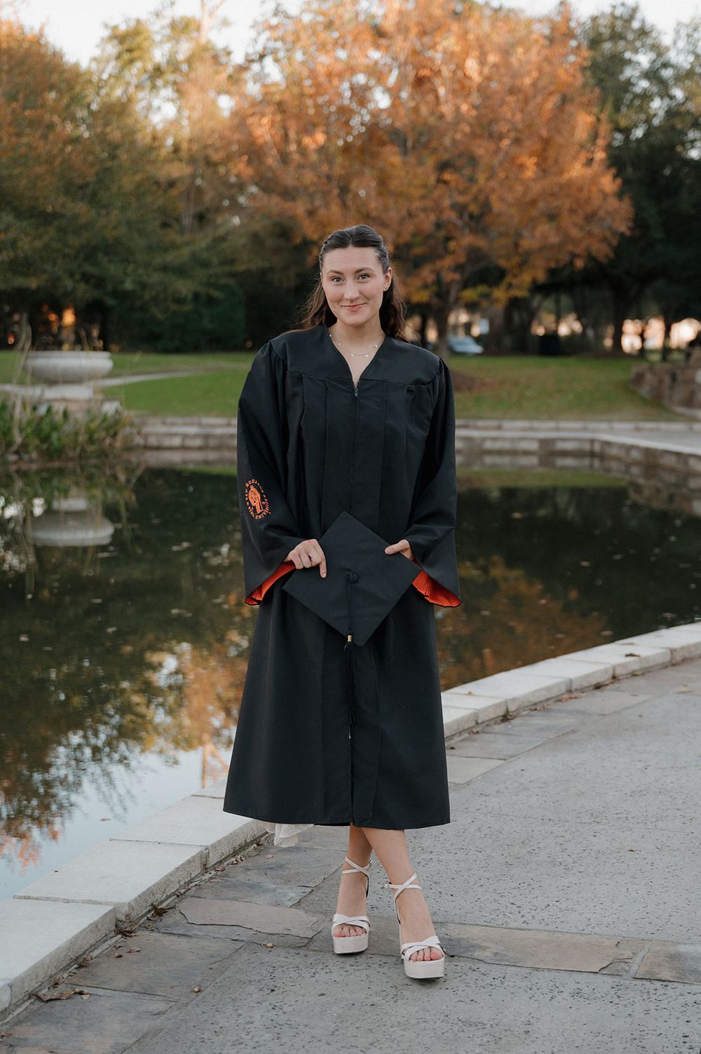 Person in a long black dress coat poses by a scenic pond with autumn trees in the background.