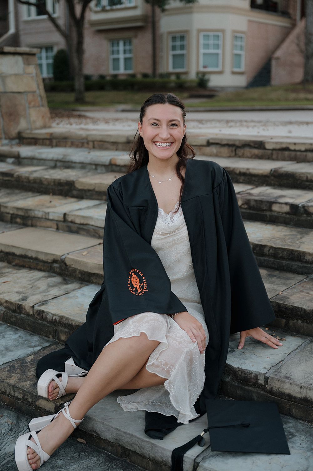 A graduate in academic regalia sits on stone steps wearing a black gown and white dress.