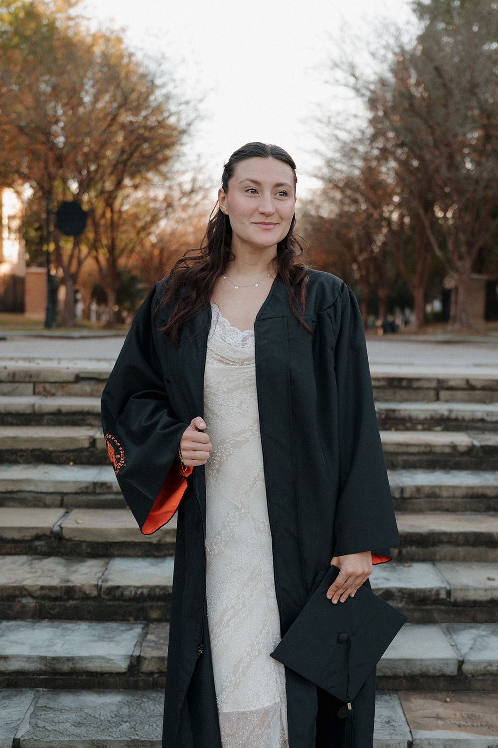 Graduate stands on stone steps in traditional black graduation gown holding cap against autumn trees in background.