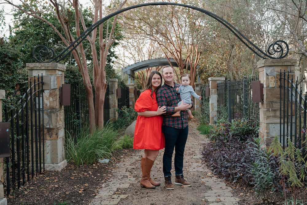 A couple stands together under an ornate metal archway on a garden path surrounded by trees and landscaping.