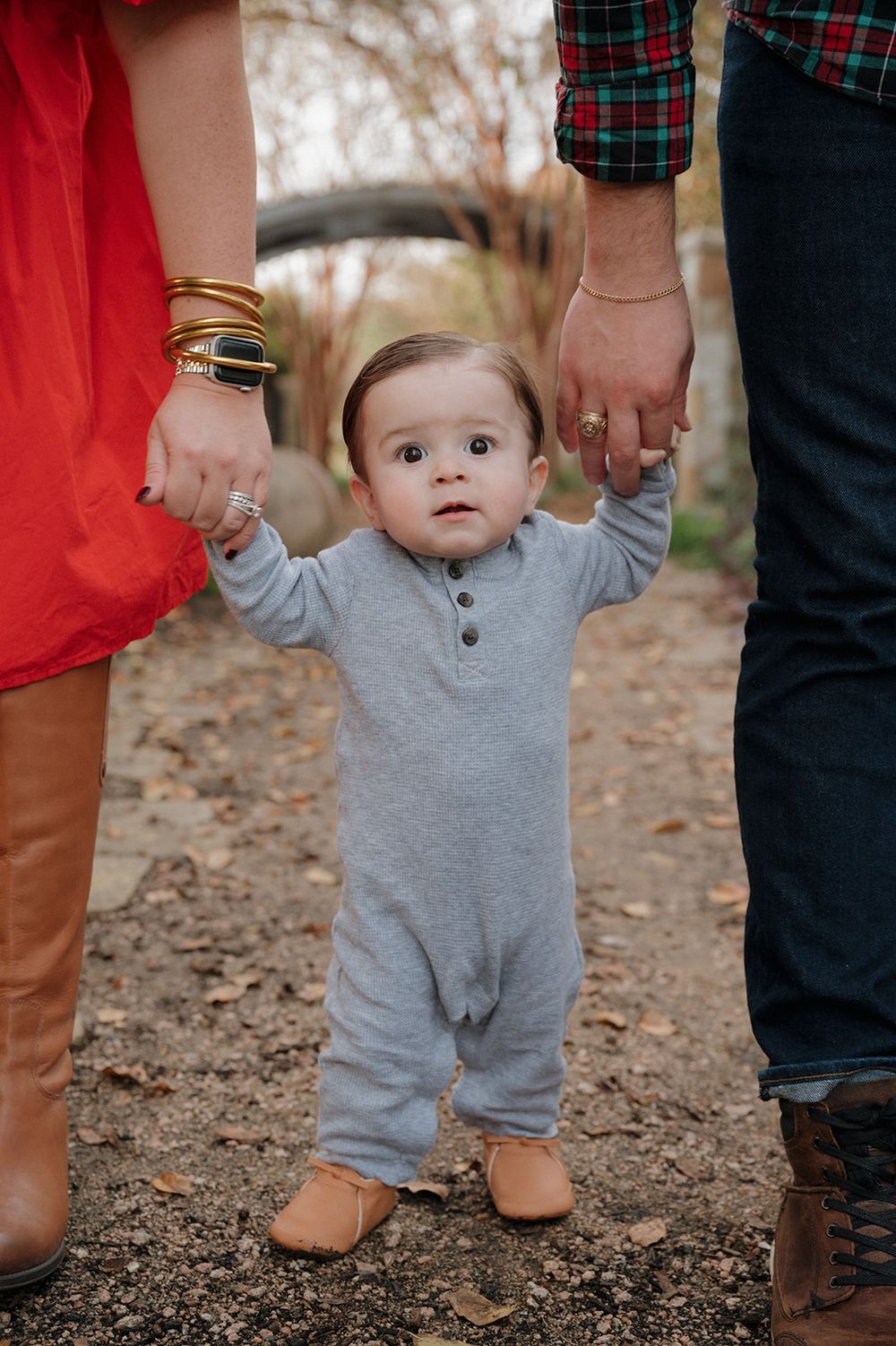 A small child in a gray onesie holding hands with parents while standing on a dirt path outdoors.
