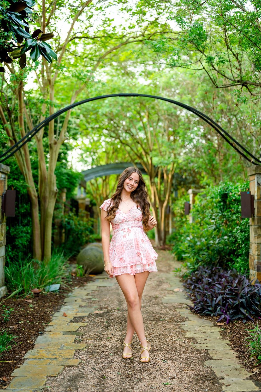 A person in a pink dress poses on a garden path beneath a green archway surrounded by lush landscaping.