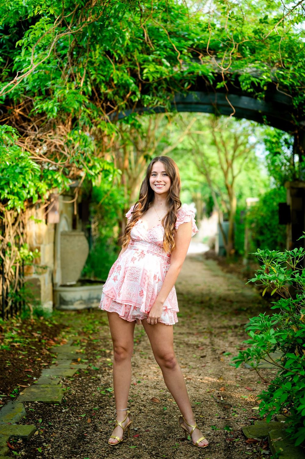 A pink dress sways in a lush garden archway path surrounded by green foliage on a sunny afternoon.