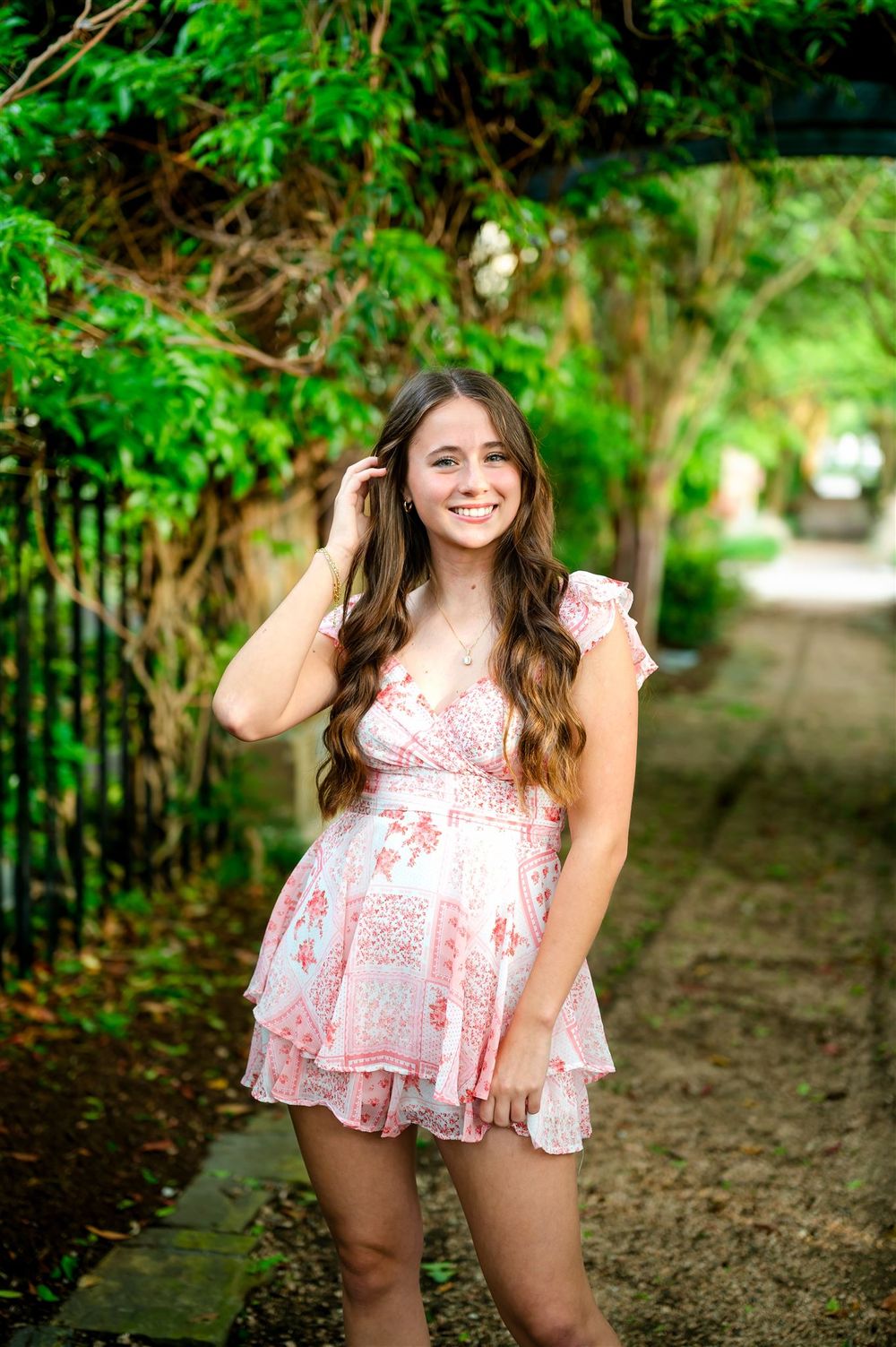 Young woman in pink floral dress smiling on garden pathway surrounded by lush greenery.