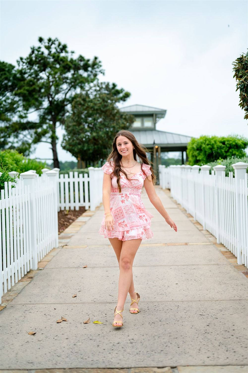 Person in a pink summer dress walks along a white picket fence-lined path towards a coastal gazebo on a sunny day.