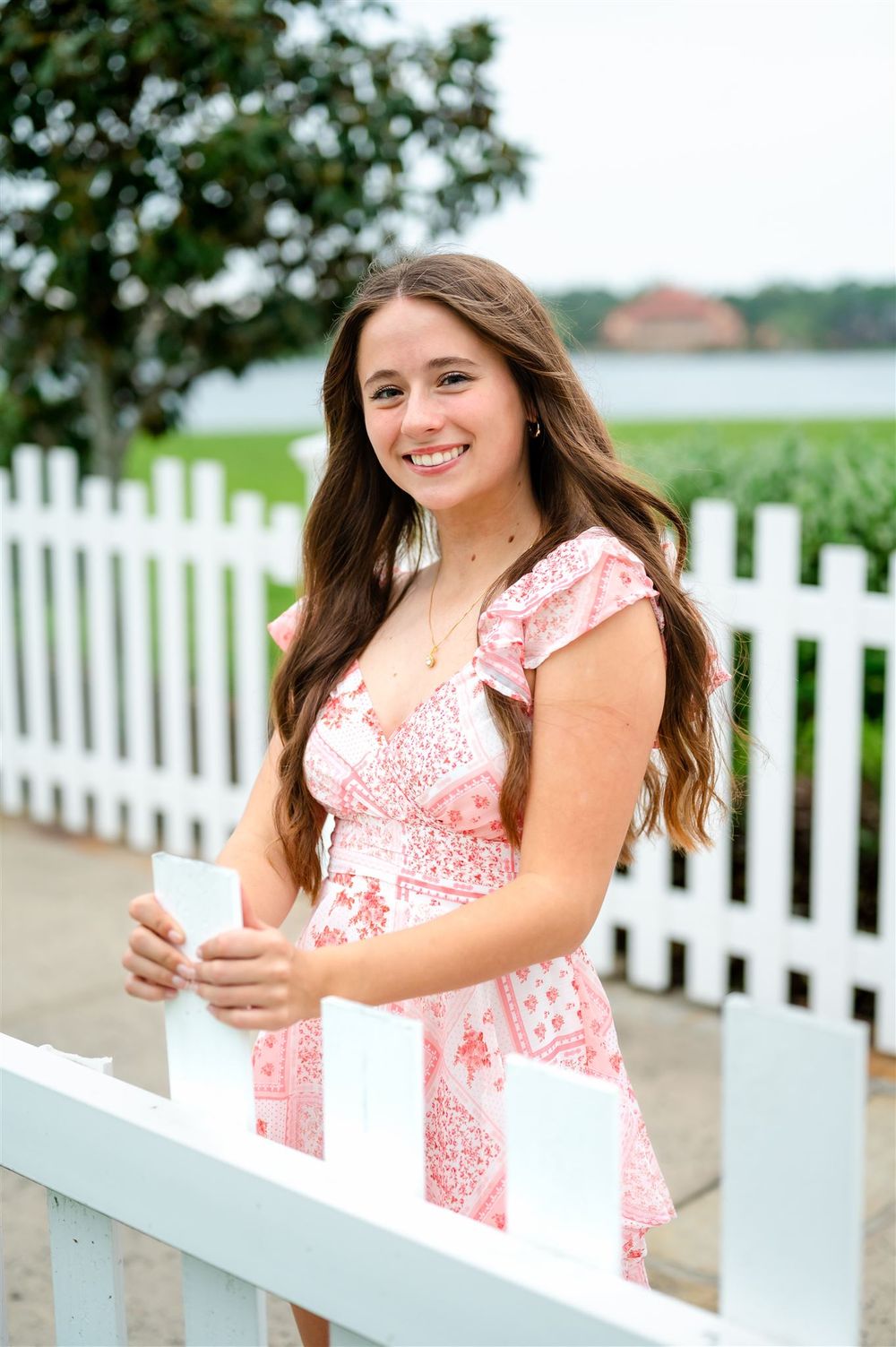 A young woman in a pink and white summer dress stands by a white picket fence near the water.