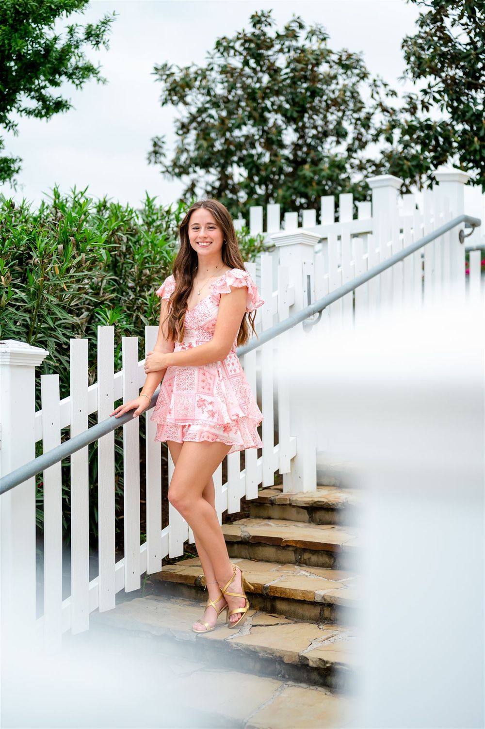 Someone in a pink floral romper poses elegantly on rustic wooden steps with white railings surrounded by greenery.