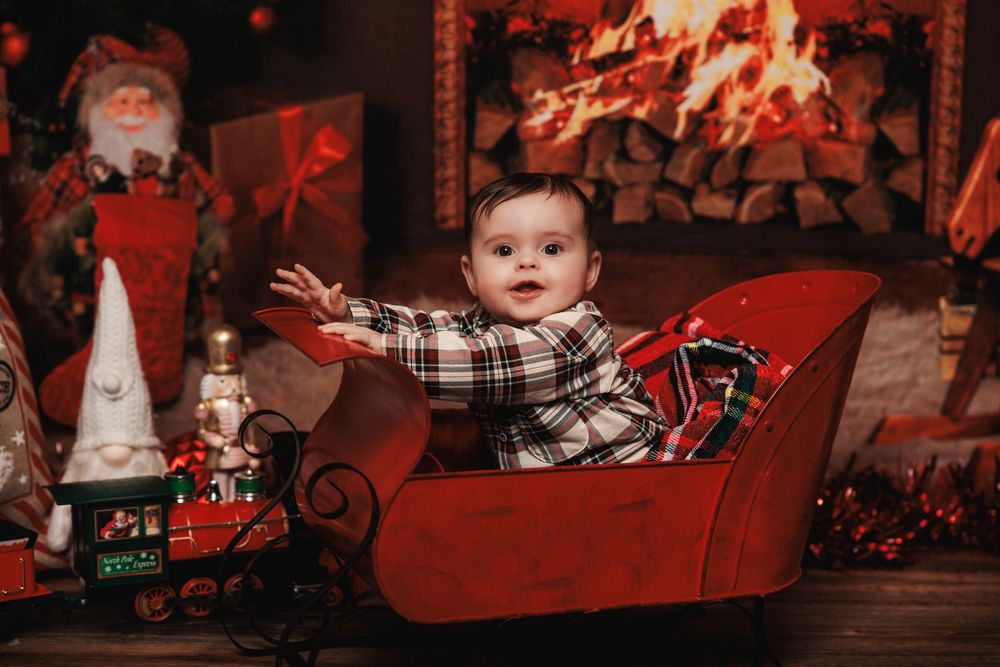 Baby in plaid clothing sits in red sleigh with Christmas decorations and fireplace in background.