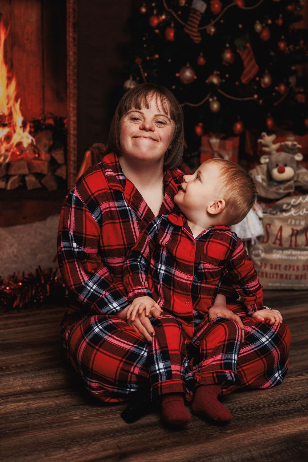 Two people in matching red plaid pajamas sit together by a fireplace with Christmas decorations in the background.