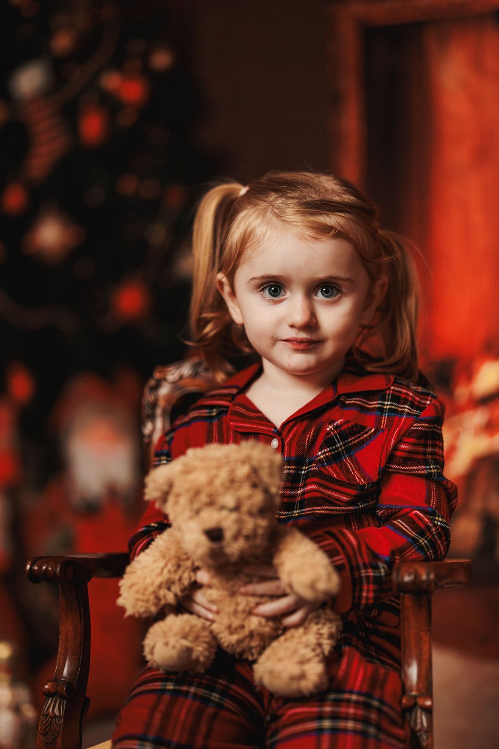 A child sits in a wooden chair holding a teddy bear while wearing a red plaid dress against a festive holiday backdrop.