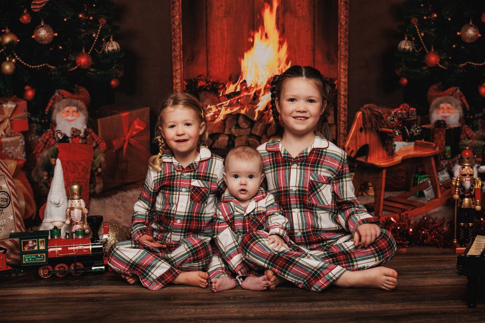 Children in matching plaid holiday pajamas sit together by a fireplace with festive Christmas decorations and a warm fire.