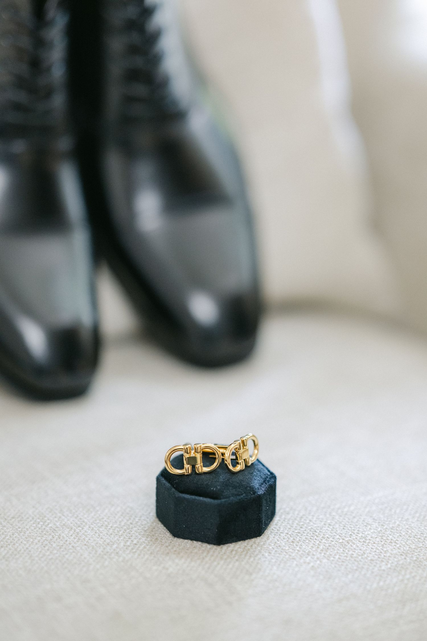 Gold wedding band resting on black velvet ring holder with dress shoes blurred in background.
