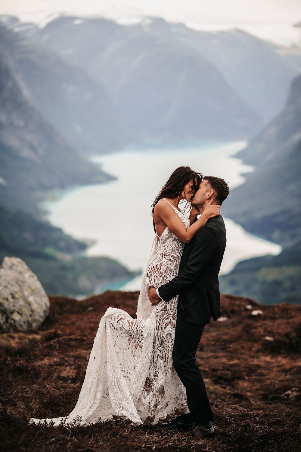 A romantic wedding couple embraces on a mountaintop overlooking a stunning glacial lake and mountain landscape.