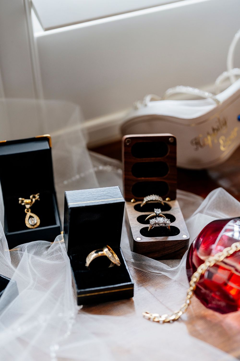 Elegant jewelry pieces including rings and necklaces displayed in black boxes on white tulle fabric next to a red perfume bottle.