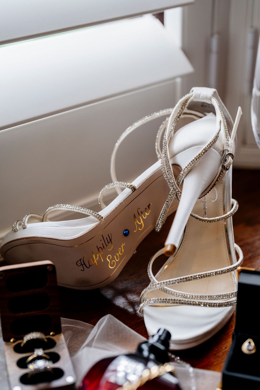 Elegant white strappy sandals with rhinestone details and high heels rest on a wooden surface near other accessories.