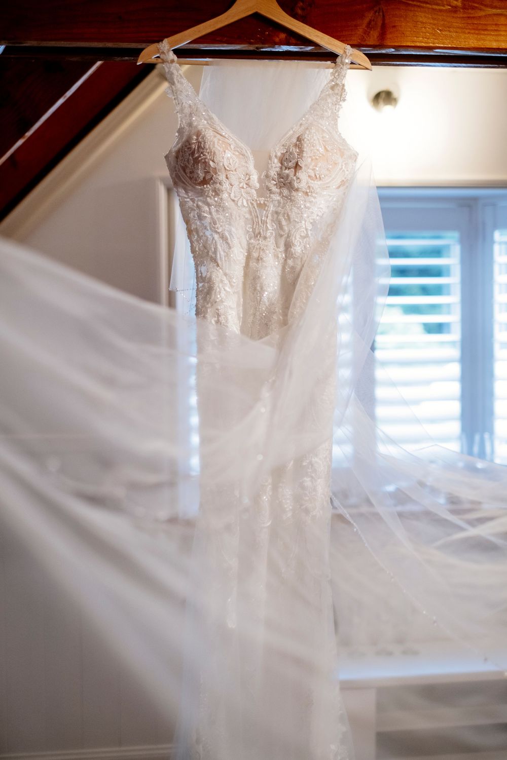 A delicate white lace wedding dress with floral details and sheer panels hangs elegantly with a flowing veil in natural light.