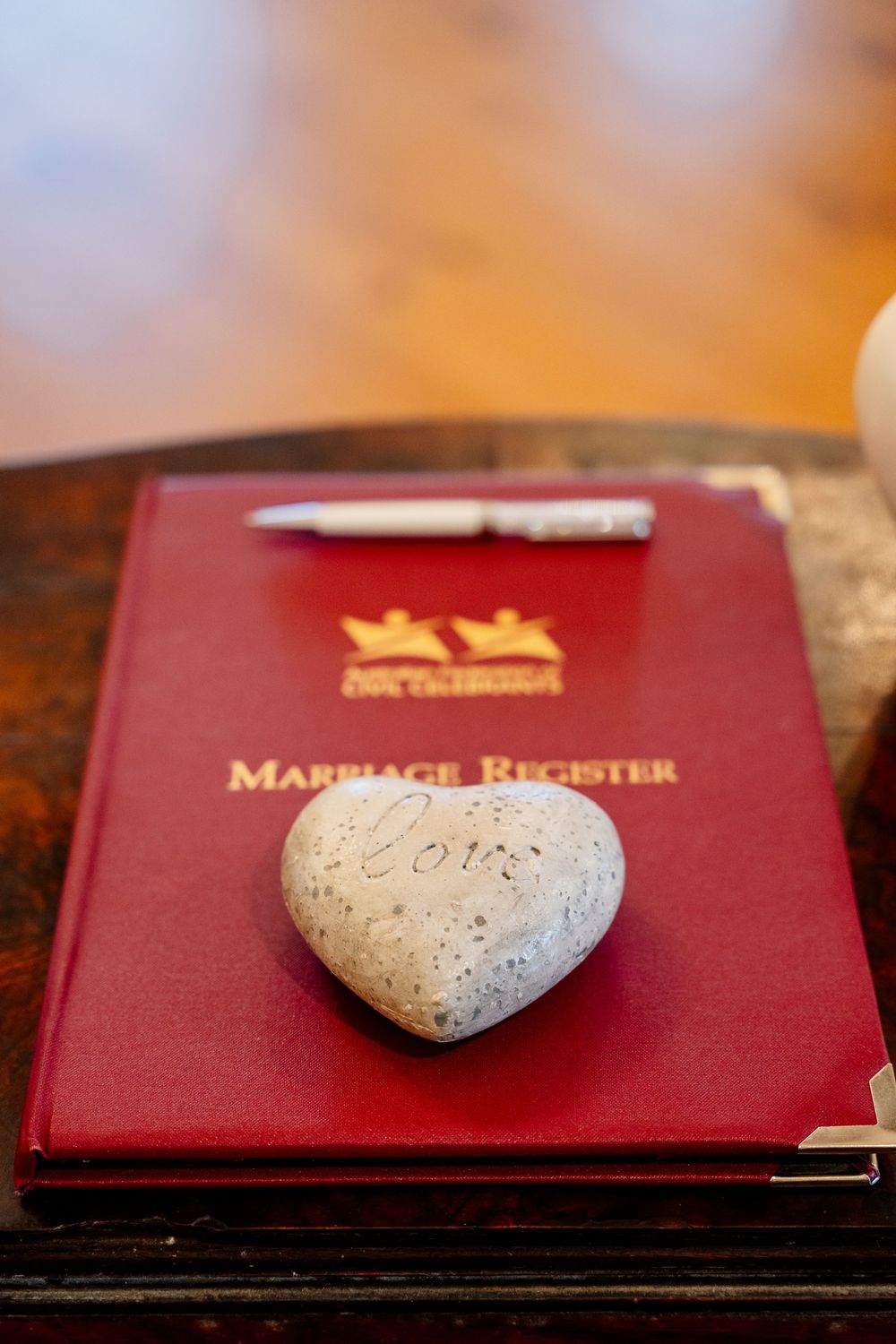 A stone heart sits atop a red marriage register book with a pen on a wooden surface.