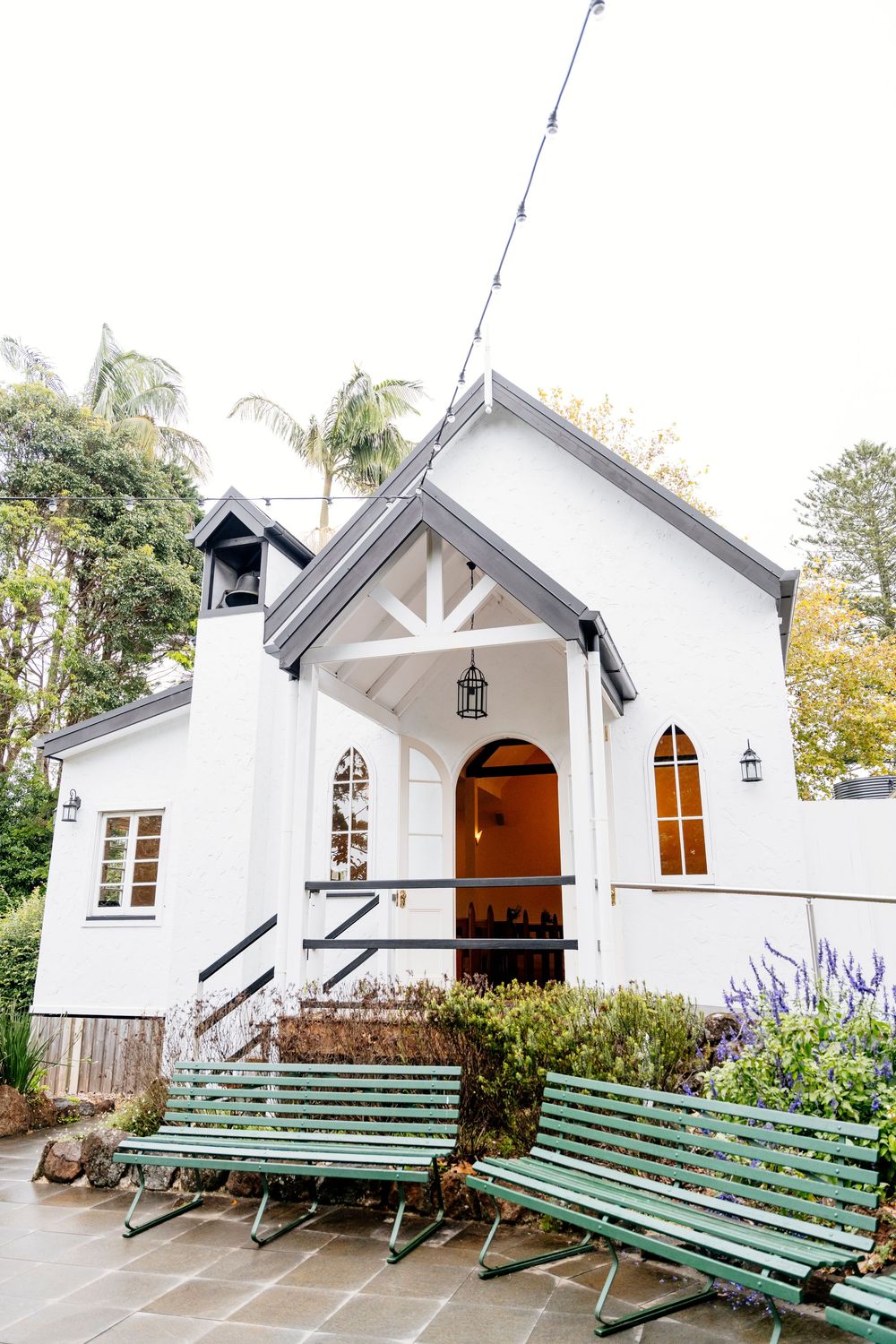 A charming white chapel with green benches and arched windows surrounded by palm trees and landscaping.