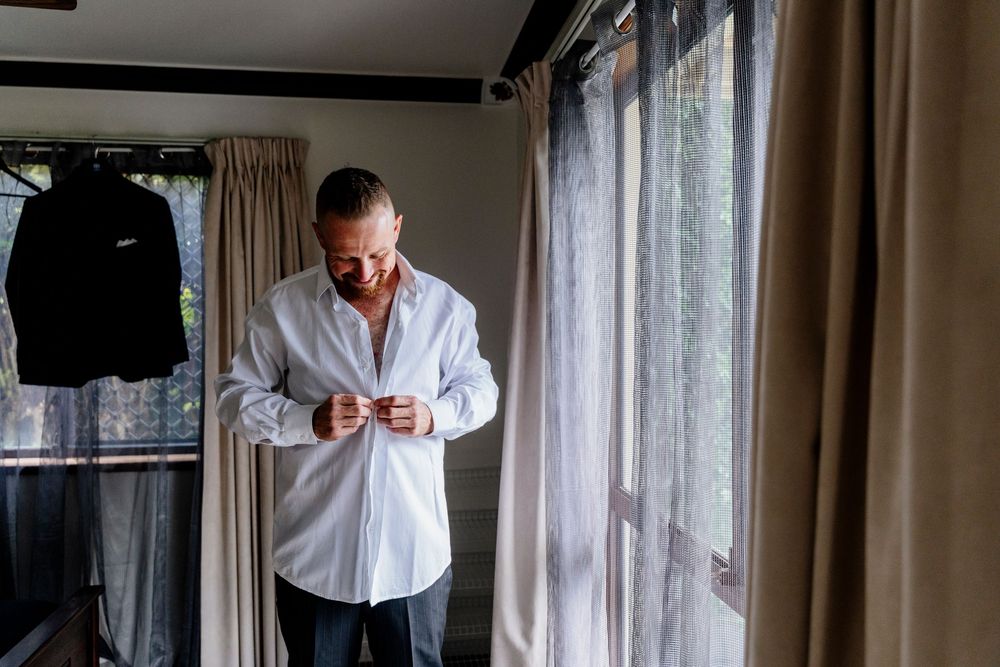 Someone in a white button-down shirt looks down at their phone while standing by curtained windows in a room.