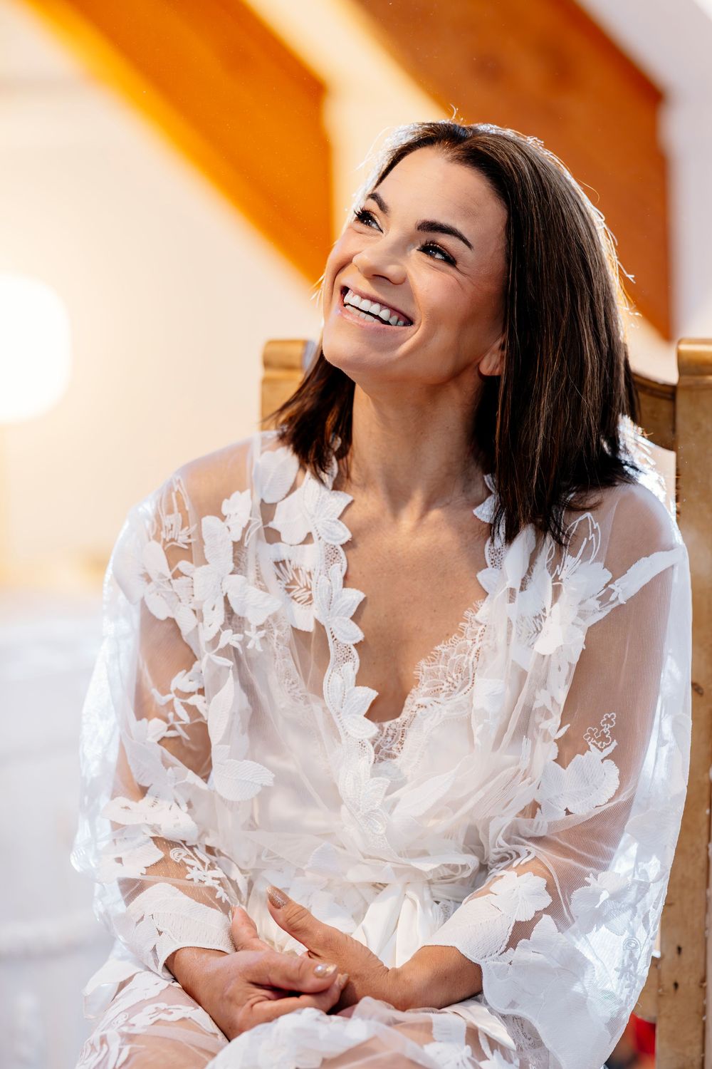 Smiling woman wearing white lace bridal robe while sitting and laughing during morning wedding preparations.