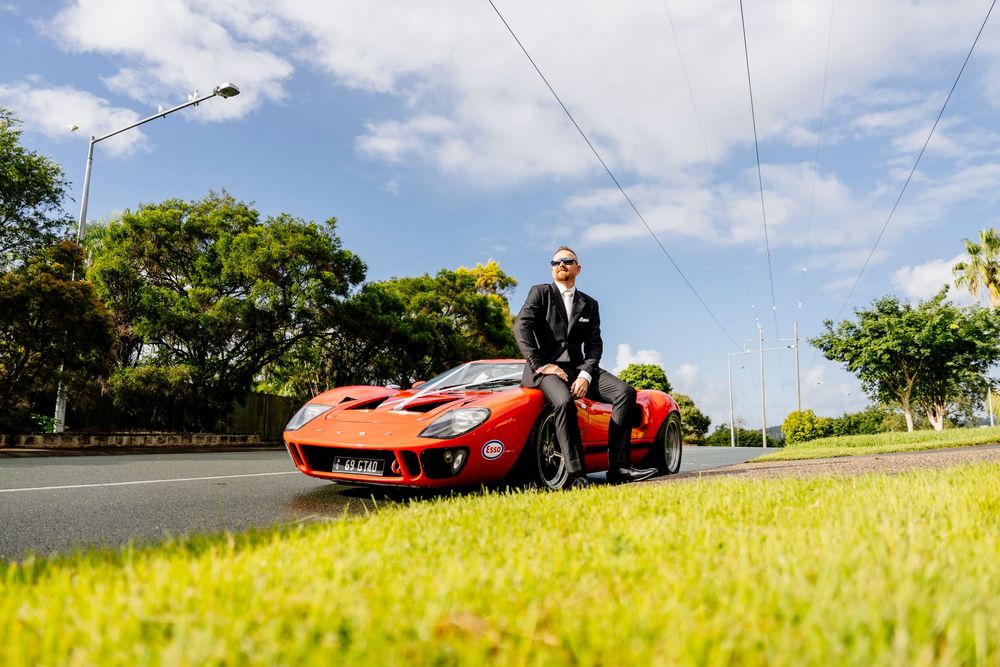 A red sports car parked on a grass verge next to a road under blue skies with white clouds.