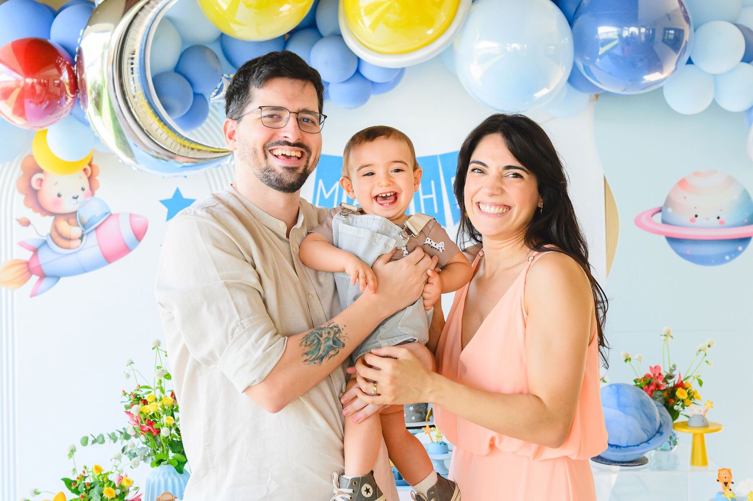 Family celebrates at baby-themed party with colorful balloon decorations and floral arrangements against pastel blue backdrop.