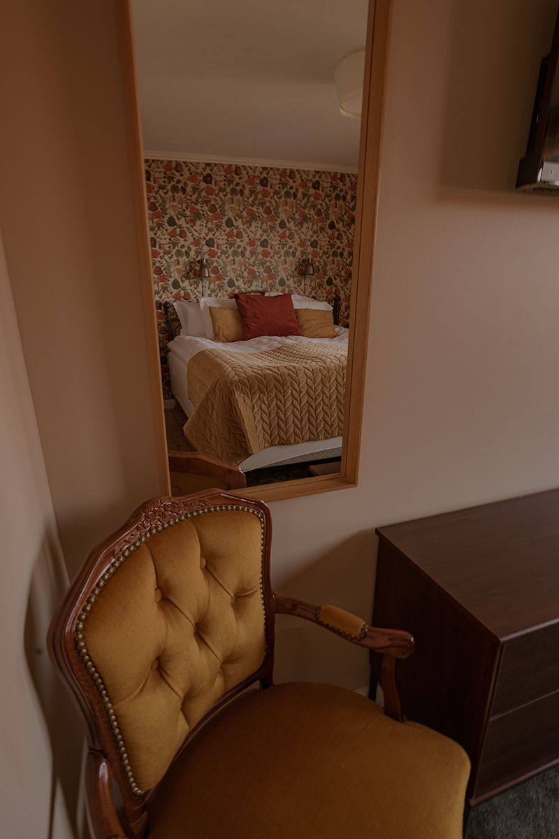 Vintage tufted beige armchair and mirror reflecting floral wallpaper and bed in cozy bedroom interior.
