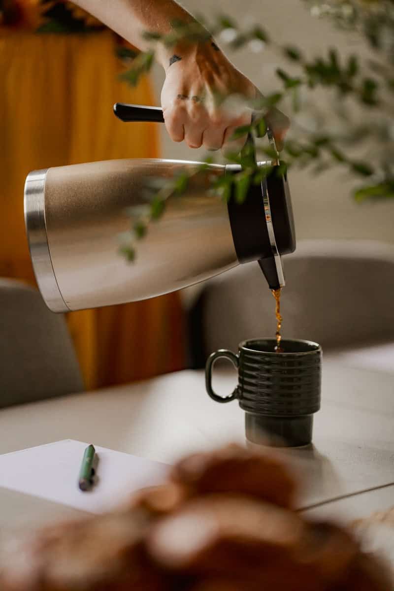 Pouring hot beverage from silver thermos into dark ceramic mug on table.
