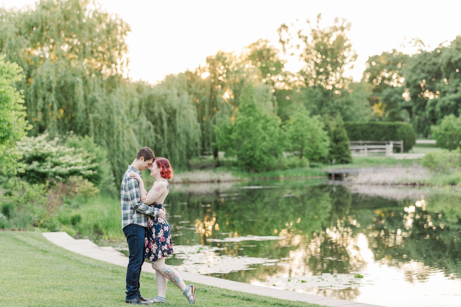 A romantic couple stands together by a serene pond surrounded by weeping willows and greenery at sunset at Cantigny Park
