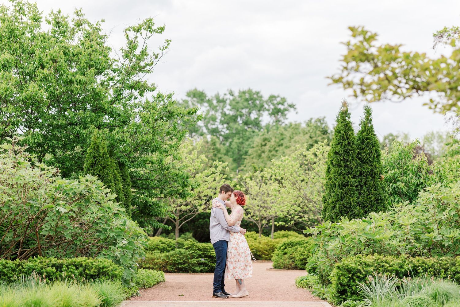 A couple shares a romantic moment in a lush garden path surrounded by green shrubbery and cypress trees at Cantigny Park