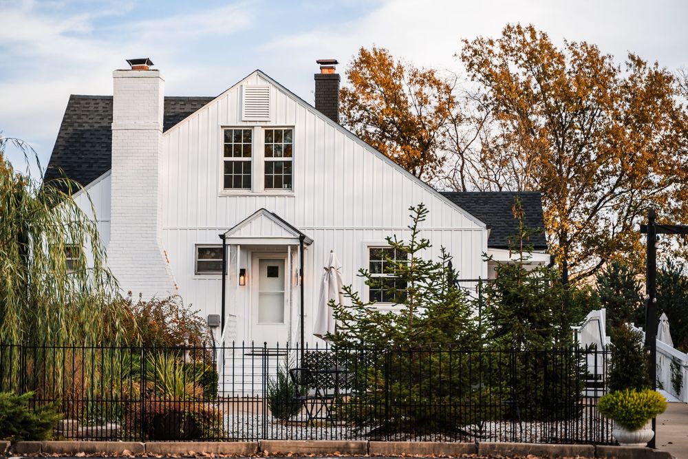 A white farmhouse style home with a tall chimney and evergreen trees behind a metal fence stands against autumn trees.