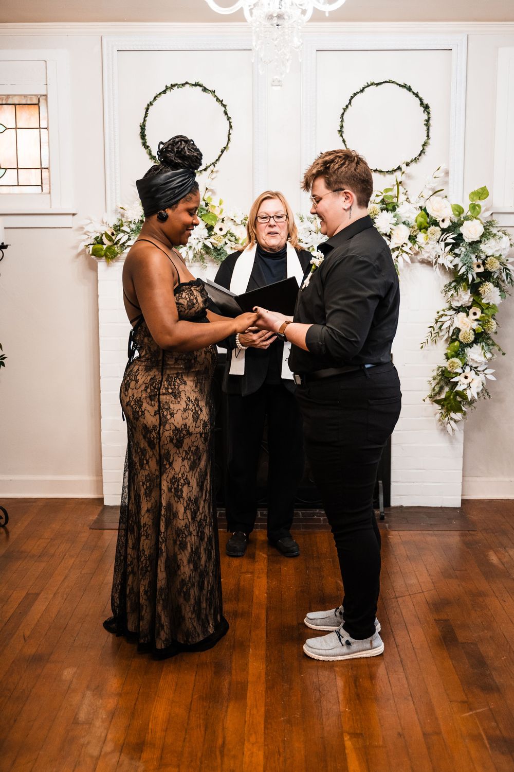 A wedding ceremony taking place in an elegant white room with floral decorations and a chandelier.