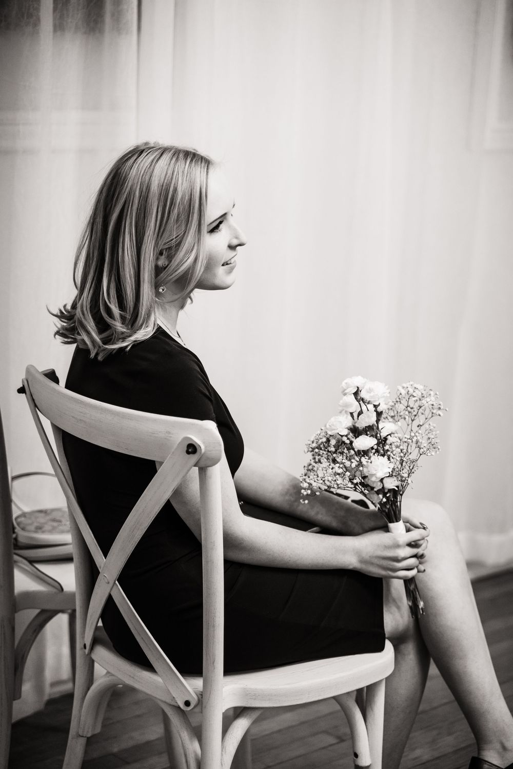 Person in black clothing holds a small bouquet of flowers while sitting on white chair.