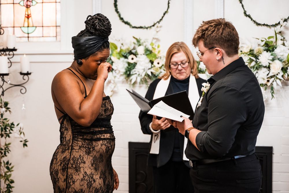 A couple shares an emotional moment during their wedding ceremony with an officiant at a venue decorated with white flowers.