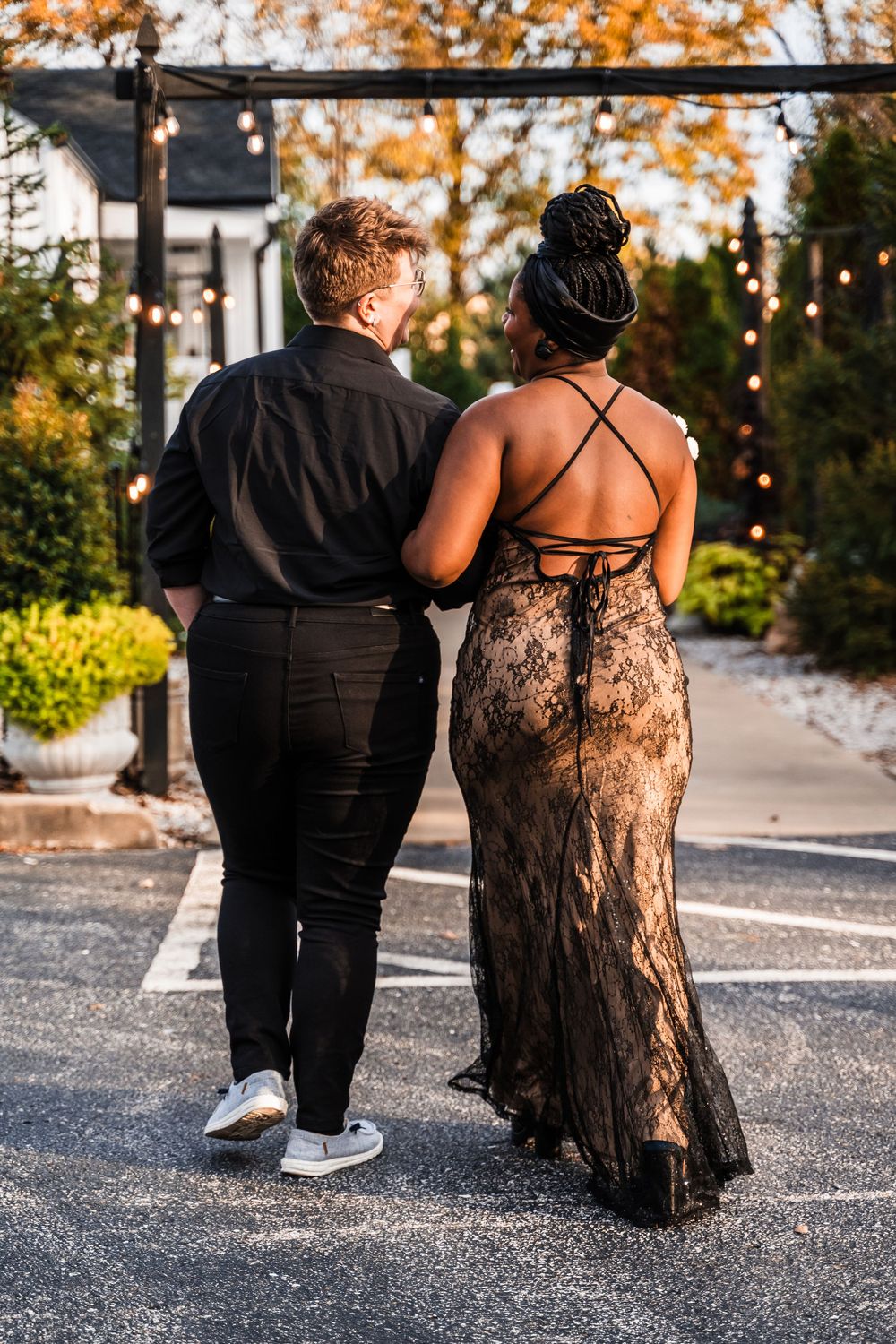 A couple walks together outdoors wearing formal attire with a black lace dress and dark suit at sunset.